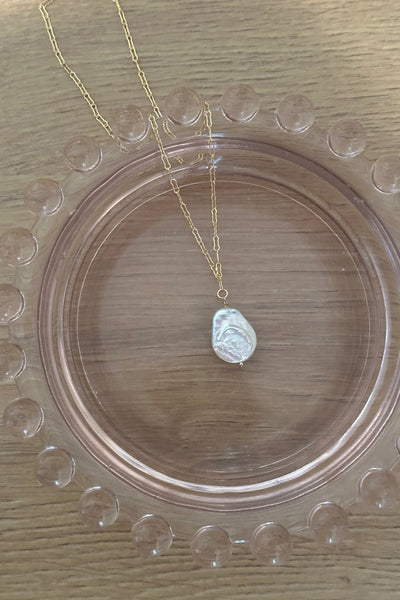 A delicate gold chain necklace with a small white textured bead pendant, displayed on a clear glass plate over a light wood grain surface.