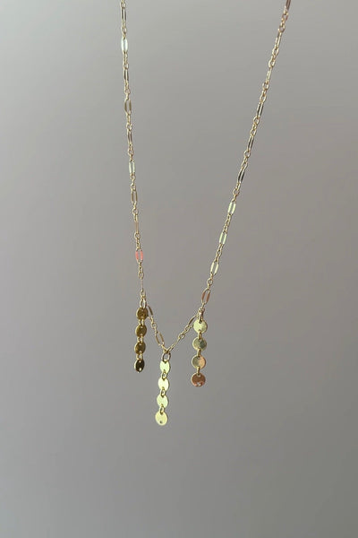 A delicate gold chain necklace with a Y-shaped pendant adorned with small, faceted gold discs, shown against a plain gray background.
