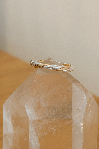 A twisted metal ring with silver and gold tones displayed on the pointed top of a clear crystal tower against a soft beige wall and light wood surface.