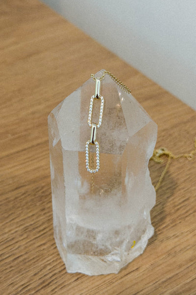 A gold-toned chain necklace with a rectangular link pendant encrusted with small clear stones, displayed on a clear crystal tower over a wooden tabletop.