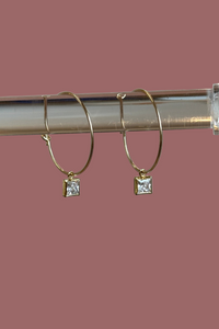 Gold hoop earrings with square gemstones on a clear stand against a neutral background