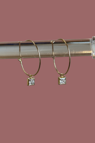 Gold hoop earrings with square gemstones on a clear stand against a neutral background
