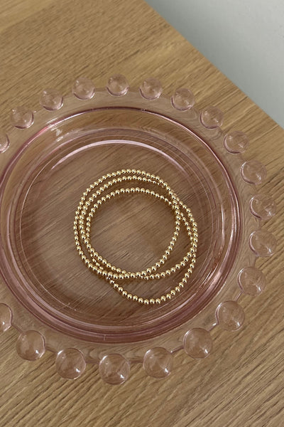 A pair of delicate gold beaded bracelets resting inside a translucent pink glass dish with a bubbly rim, placed on a light wood tabletop.
