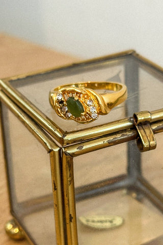 Gold ring with a green gemstone in a glass display box on a wooden surface.