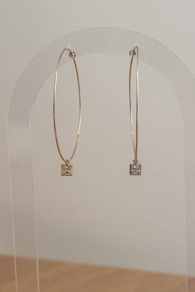 Gold hoop earrings with square gemstones on a clear stand against a white background