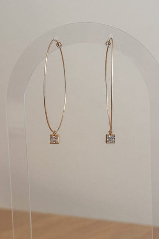Gold hoop earrings with square gemstones on a clear stand against a white background